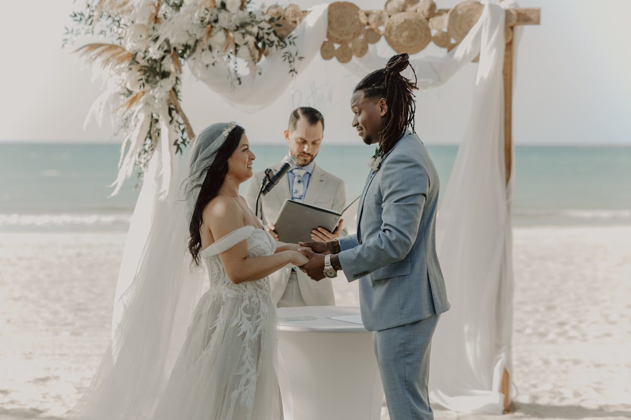 A romantic beach wedding ceremony with a bride and groom holding hands, guided by an officiant.